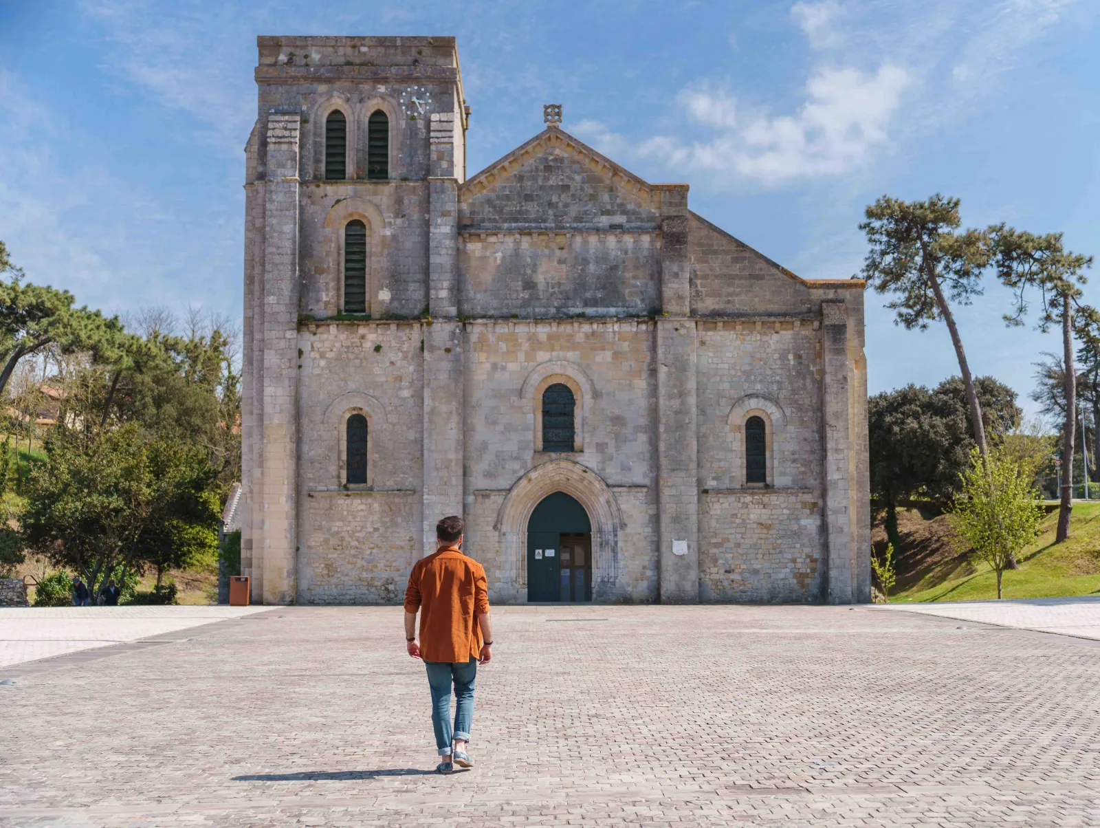 A visitor in an orange shirt standing before the Basilica of Notre-Dame-de-la-fin-des-Terres in Bordeaux, a Romanesque stone building with a square tower and austere facade.