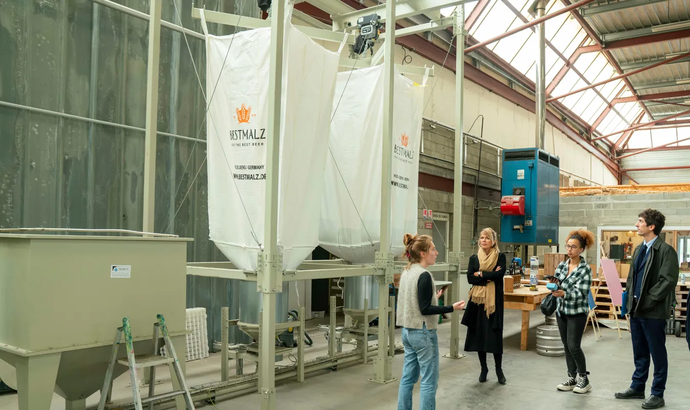 Guided tour of the Bestmalz brewery in Bordeaux with visitors observing fermentation tanks and brewing equipment in an industrial warehouse.