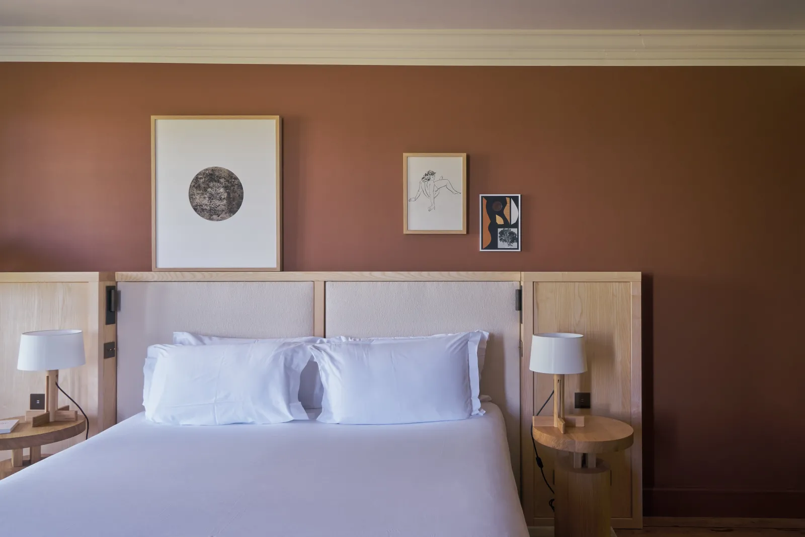 Elegant hotel bedroom with terracotta walls, king-size bed with gray upholstered headboard, wooden nightstands, white lamps, and three framed artworks above the bed.