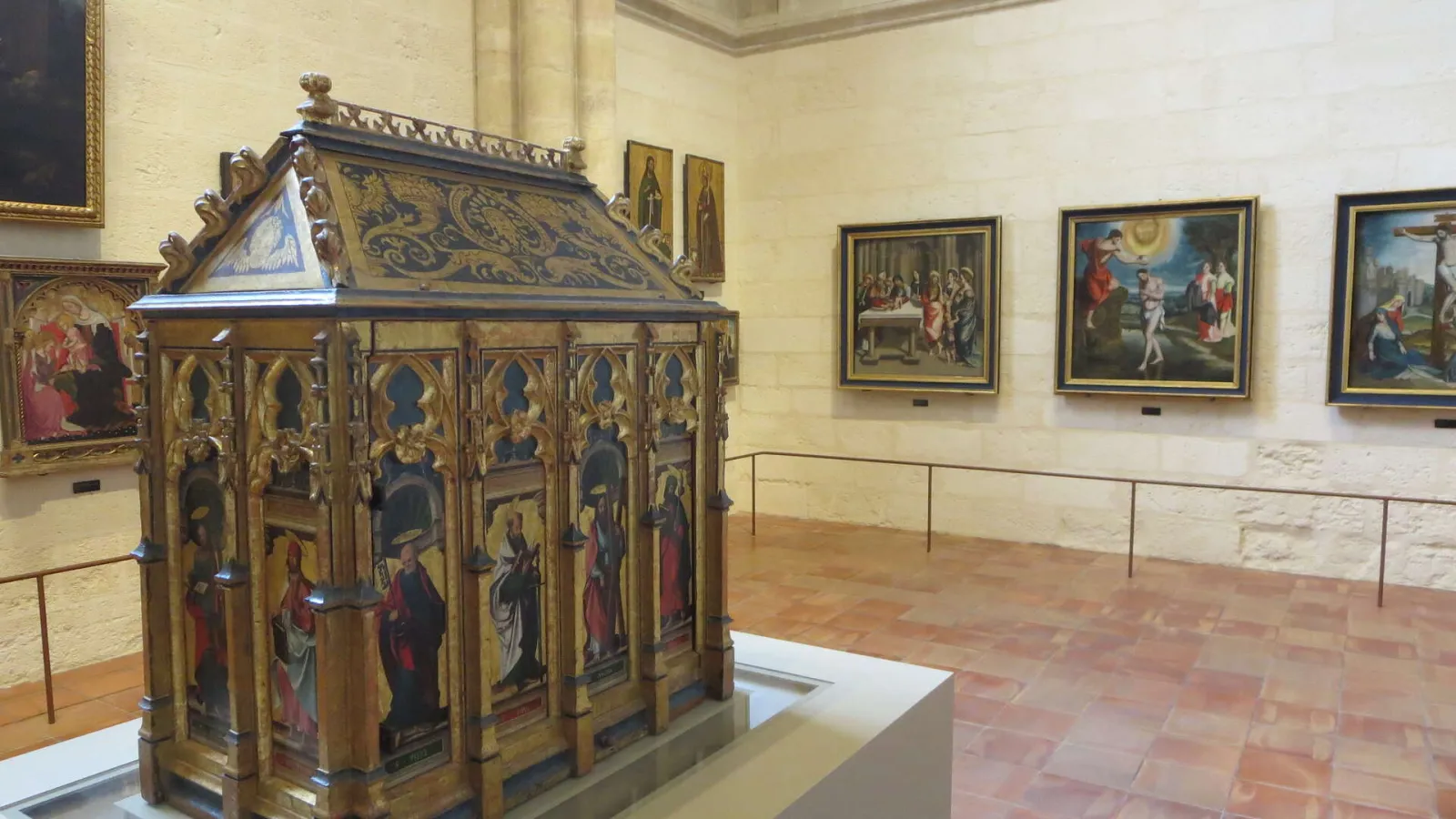 Interior of an art gallery in Bordeaux Cathedral featuring a gilded Gothic altarpiece in the foreground with painted panels depicting saints, and multiple religious paintings displayed on the walls in the background.
