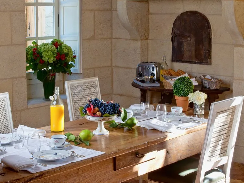 Elegant dining table set with fresh fruits, flowers and water glasses, in a stone-walled room with white chairs and floral decorations.