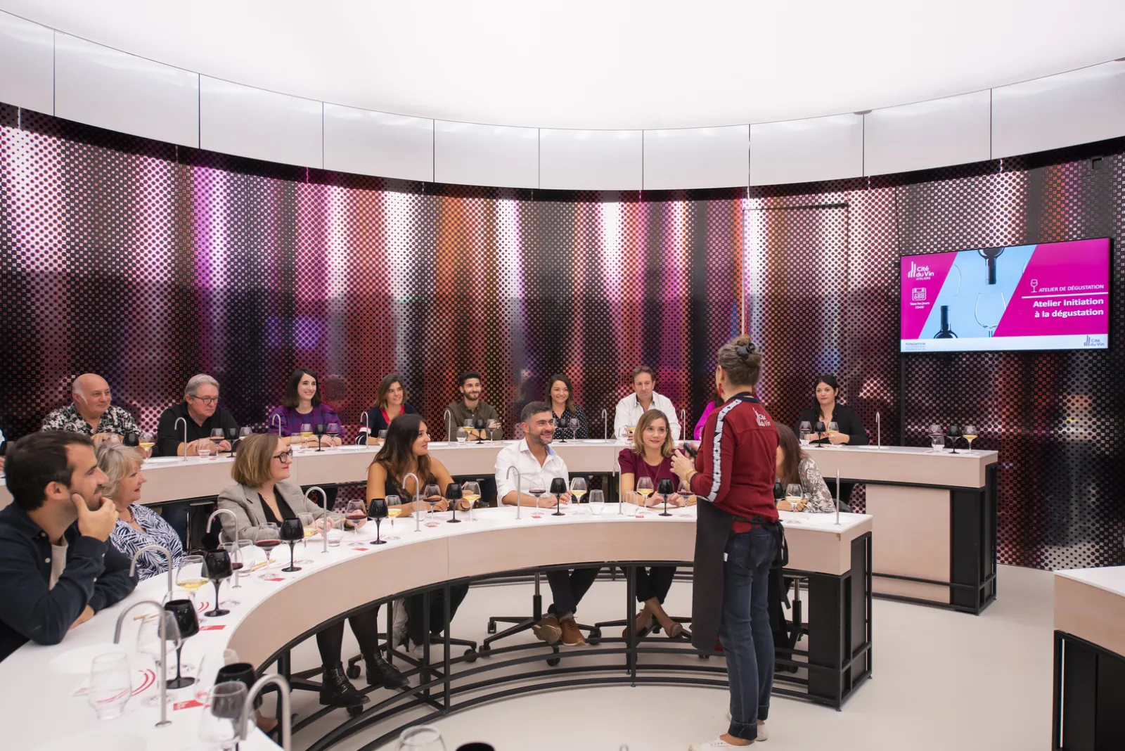 Professional meeting in a modern circular conference room at Bordeaux's Cité du Vin, with participants seated around curved tables, a pink display screen in the background, and illuminated wall panels.
