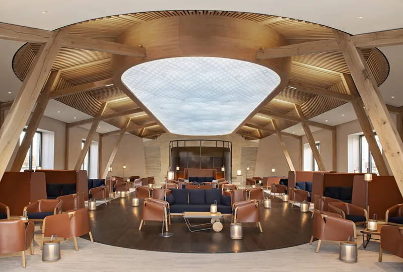 Modern interior of a lounge at Château de Ferrand in Bordeaux, featuring a light wood geometric ceiling, a large skylight, cognac leather armchairs symmetrically arranged, and minimalist contemporary architecture.