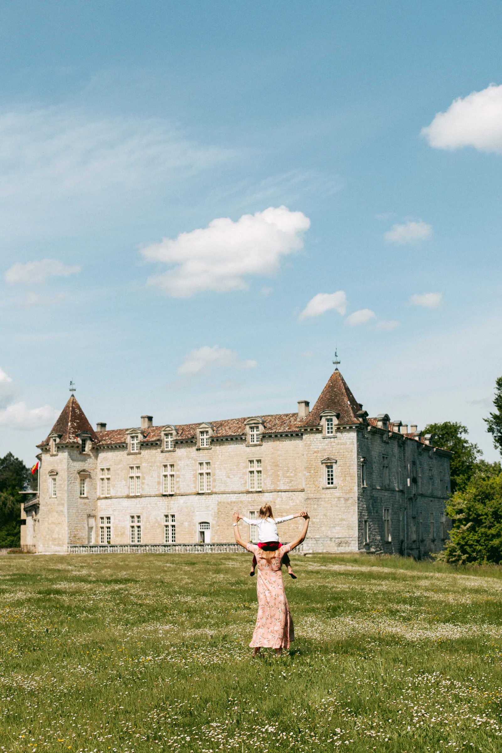 A woman in a pink dress with raised arms faces the Royal Castle of Cazeneuve, standing in a flowery meadow under a blue sky with white clouds.