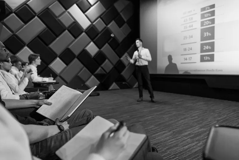Professional presentation in a Bordeaux conference room with a speaker addressing an audience holding documents, in front of a screen displaying statistical data and a geometric wall backdrop.
