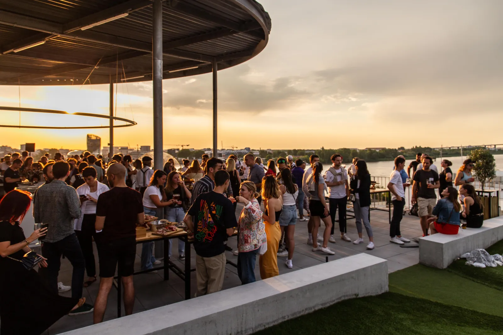 Crowd of people socializing at sunset on a covered terrace overlooking the Garonne river in Bordeaux, with city views in the background.