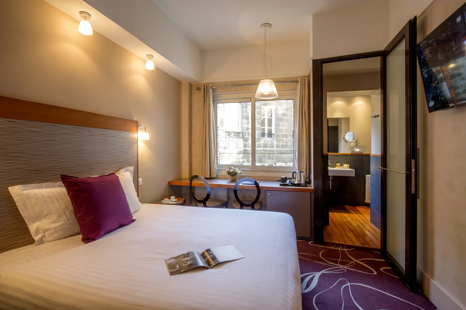 Elegant hotel room featuring a queen bed with textured headboard, work desk, ensuite bathroom, and views over Bordeaux rooftops