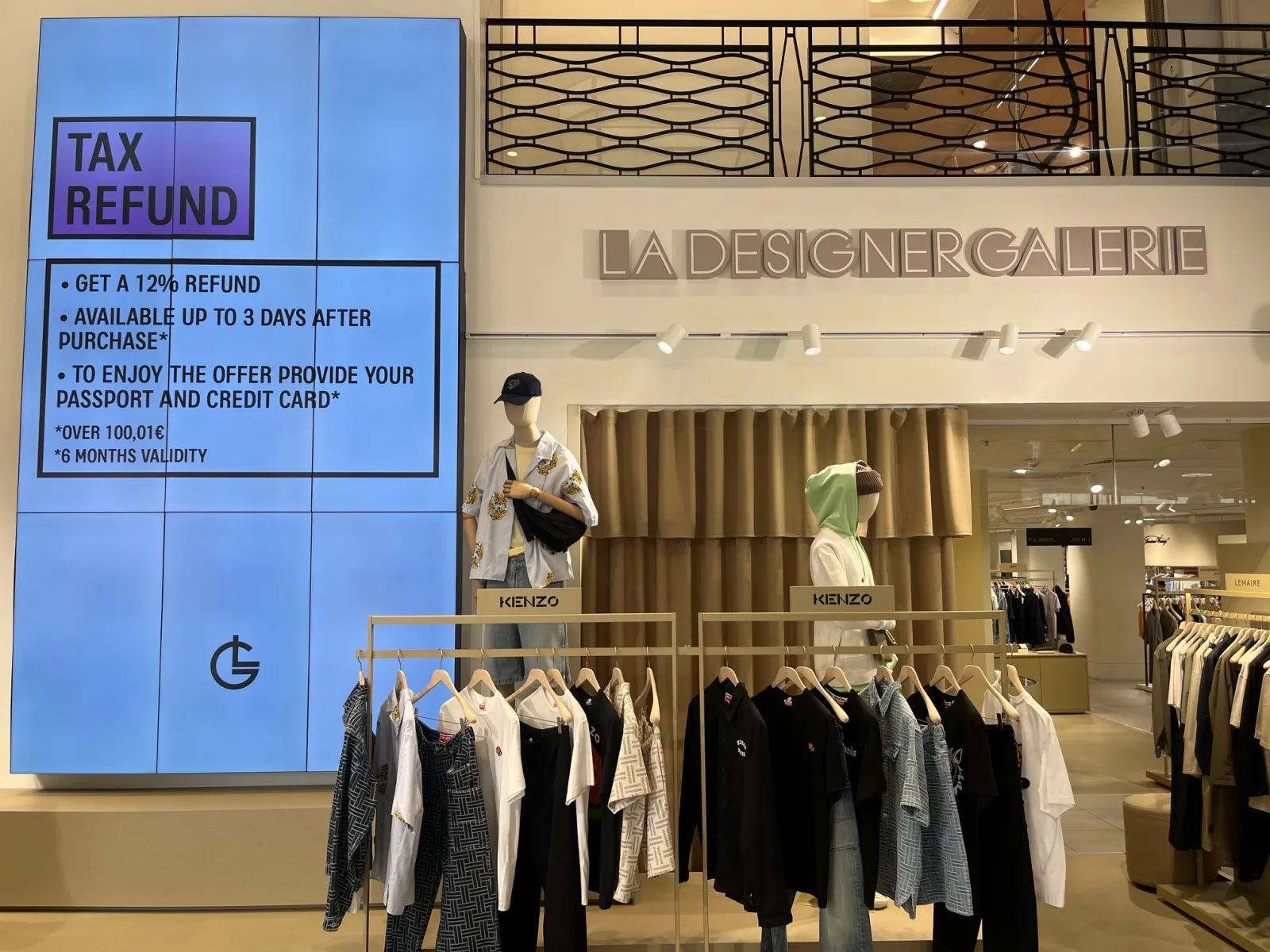 Storefront of La Designer Galerie at Galeries Lafayette in Bordeaux featuring a blue advertising sign offering 12% tax refund, Kenzo clothing displays on mannequins, and clothing racks with black and white garments.