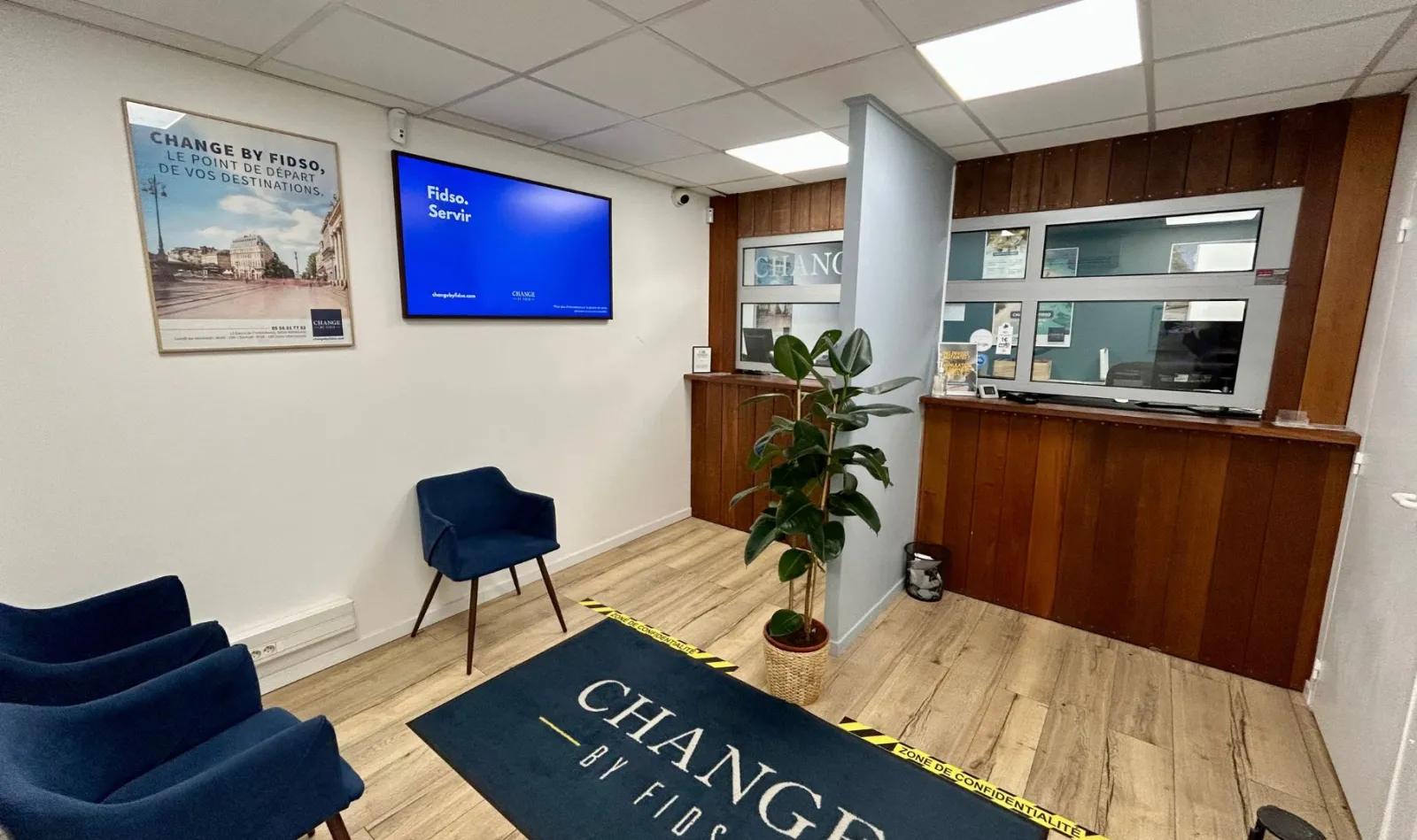 Interior of a Fidso Change exchange office in Bordeaux featuring a service counter, blue chairs, digital display screen, potted plant, and yellow and black floor signage