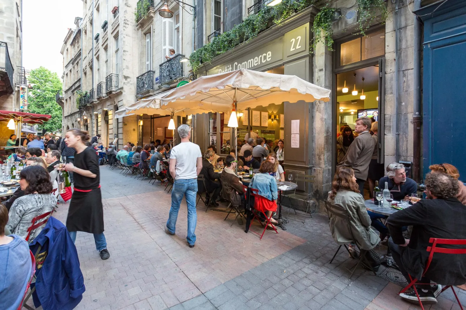 Busy café terrace on a paved street in Bordeaux, with diners seated under white and yellow parasols, stone buildings and shops in the background.