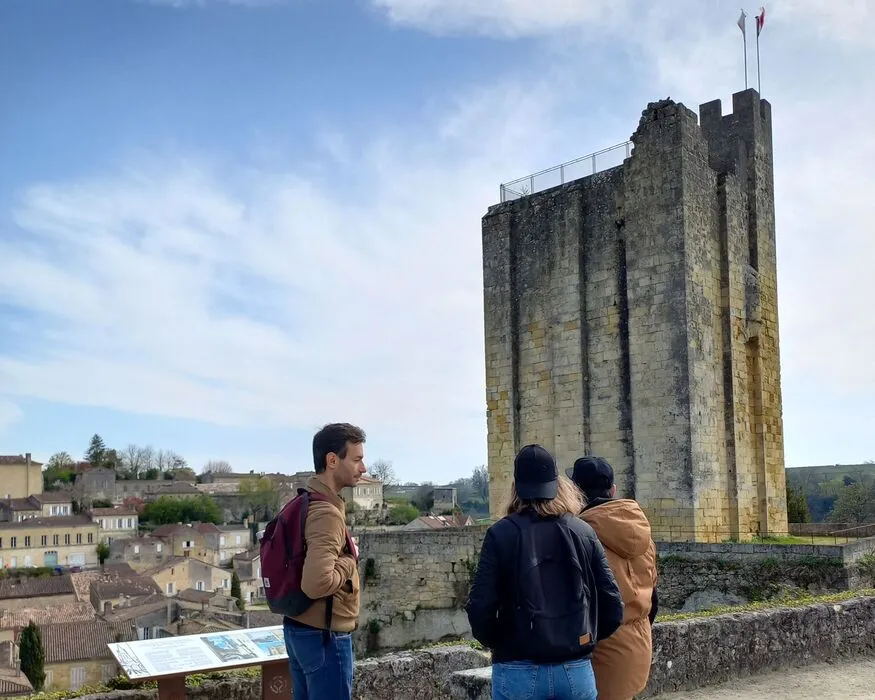 Three visitors looking at the Tour de Vesone, an ancient Roman tower in Bordeaux, with the historic city visible in the background and an information panel in the foreground.