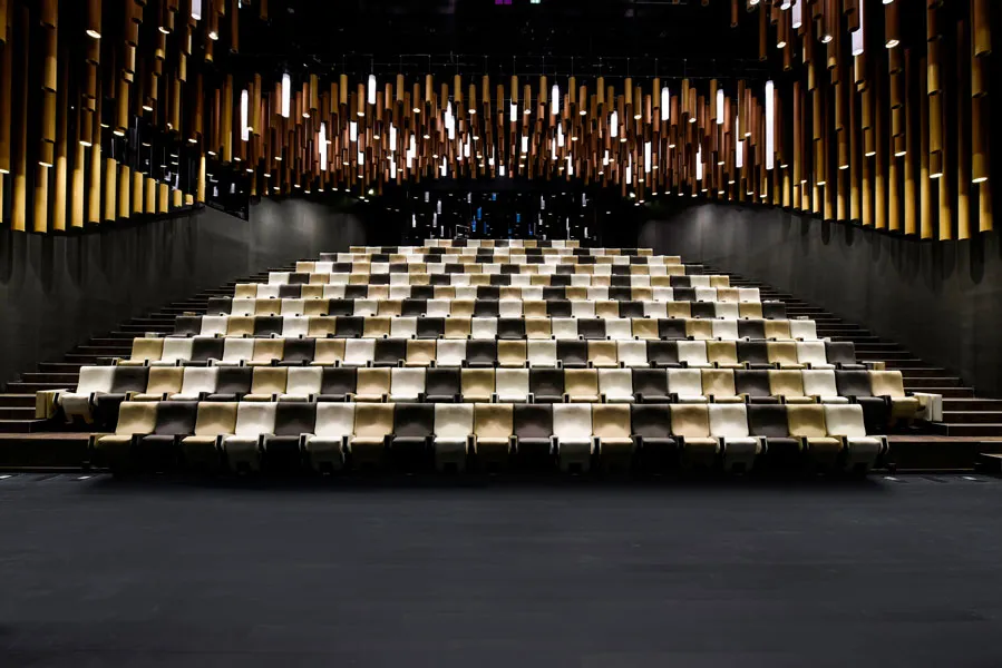 Interior of the Thomas Jefferson Auditorium at La Cité du Vin in Bordeaux, featuring tiered seating in beige and black tones, with an illuminated ceiling featuring golden and white light panels.