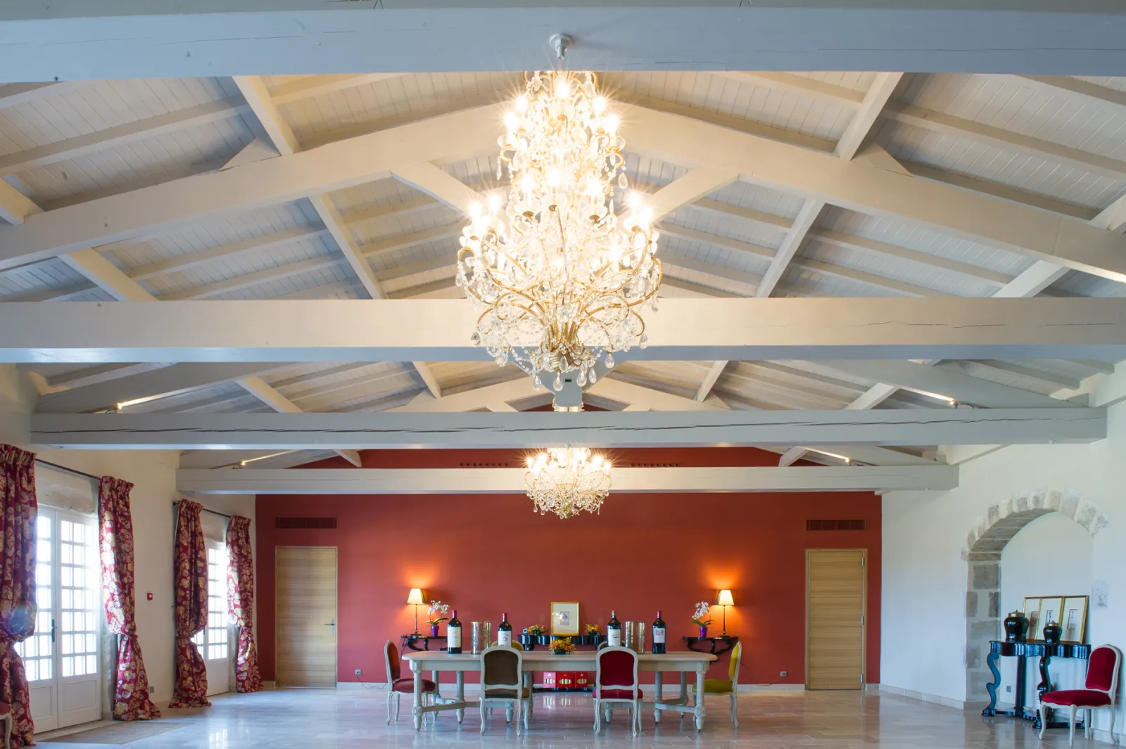 Elegant grand hall of Château de Rayne Vigneau in Bordeaux featuring exposed beam ceiling, crystal chandelier, deep red accent wall, tasting furniture and large windows.