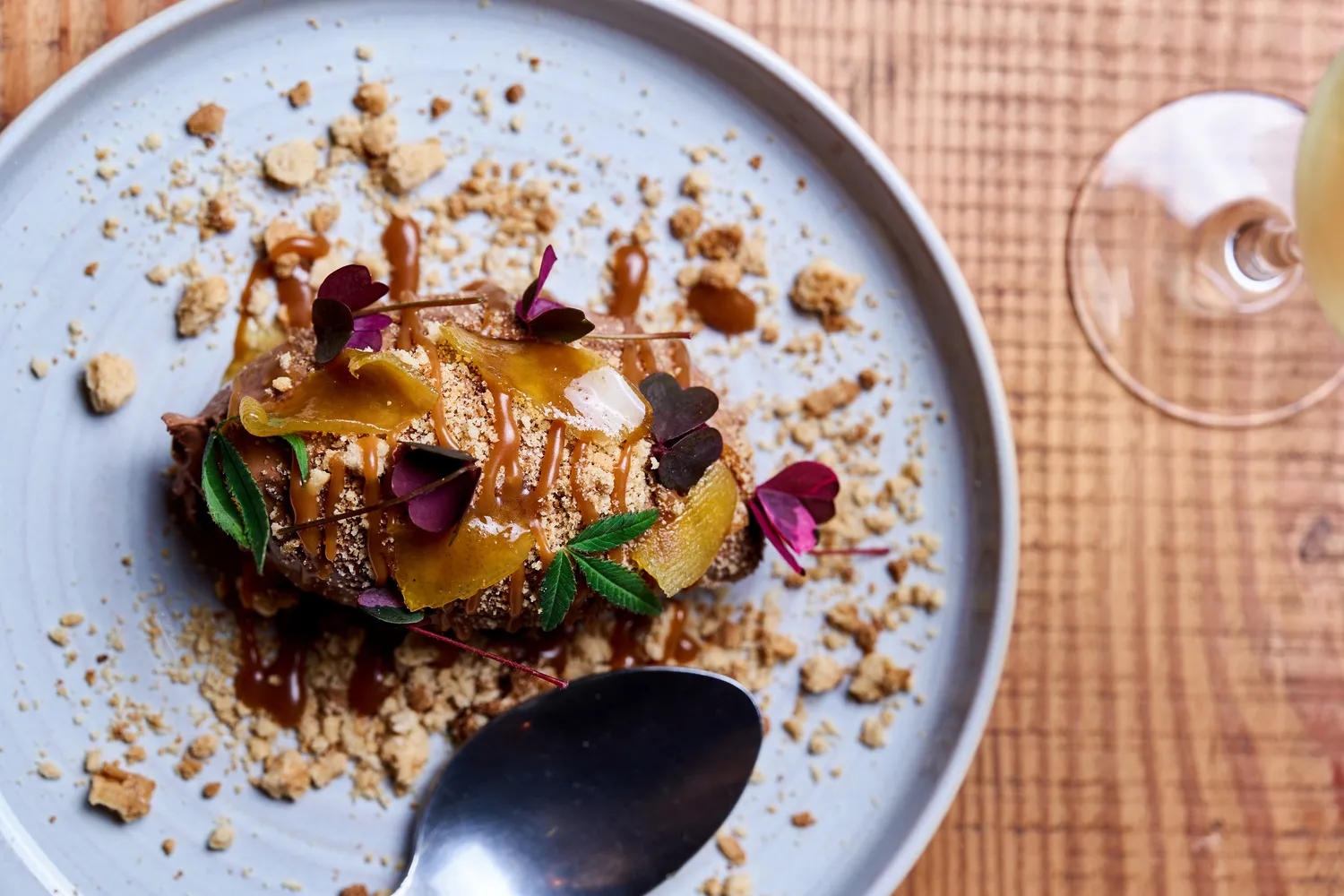 Fine dining plate featuring an elegant chocolate dessert topped with candied fruits, purple edible flowers, and cookie crumble, served with a glass of white wine.