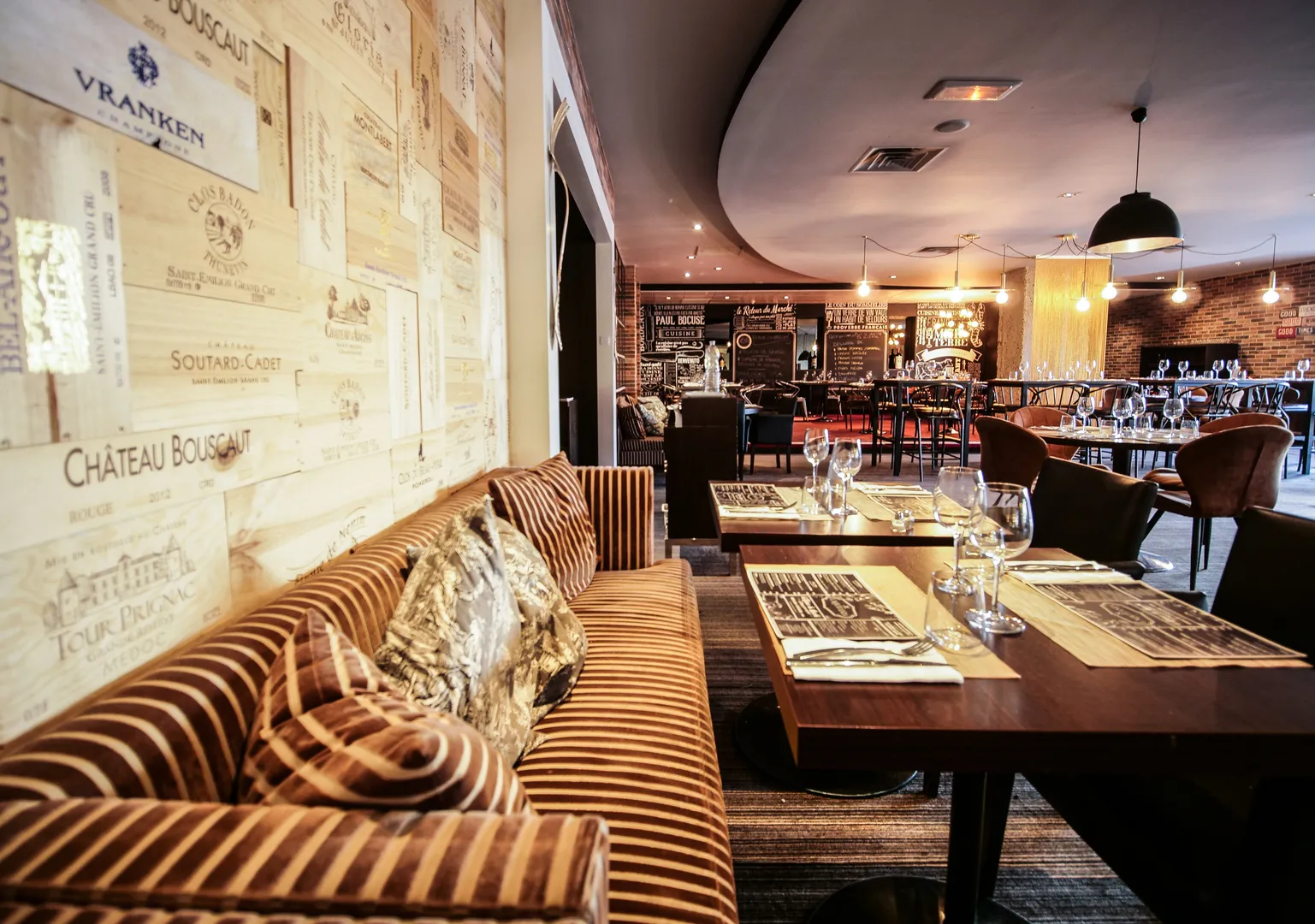 Elegant restaurant dining room at Pullman Bordeaux Lac hotel featuring a striped golden sofa, set tables, bar counter, and Bordeaux wine label wall decoration