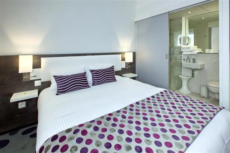 Modern hotel room featuring a double bed with white linens, purple striped pillows, geometric patterned bedspread in purple and grey, dark wood headboard with wall sconces, and an ensuite bathroom with walk-in shower.
