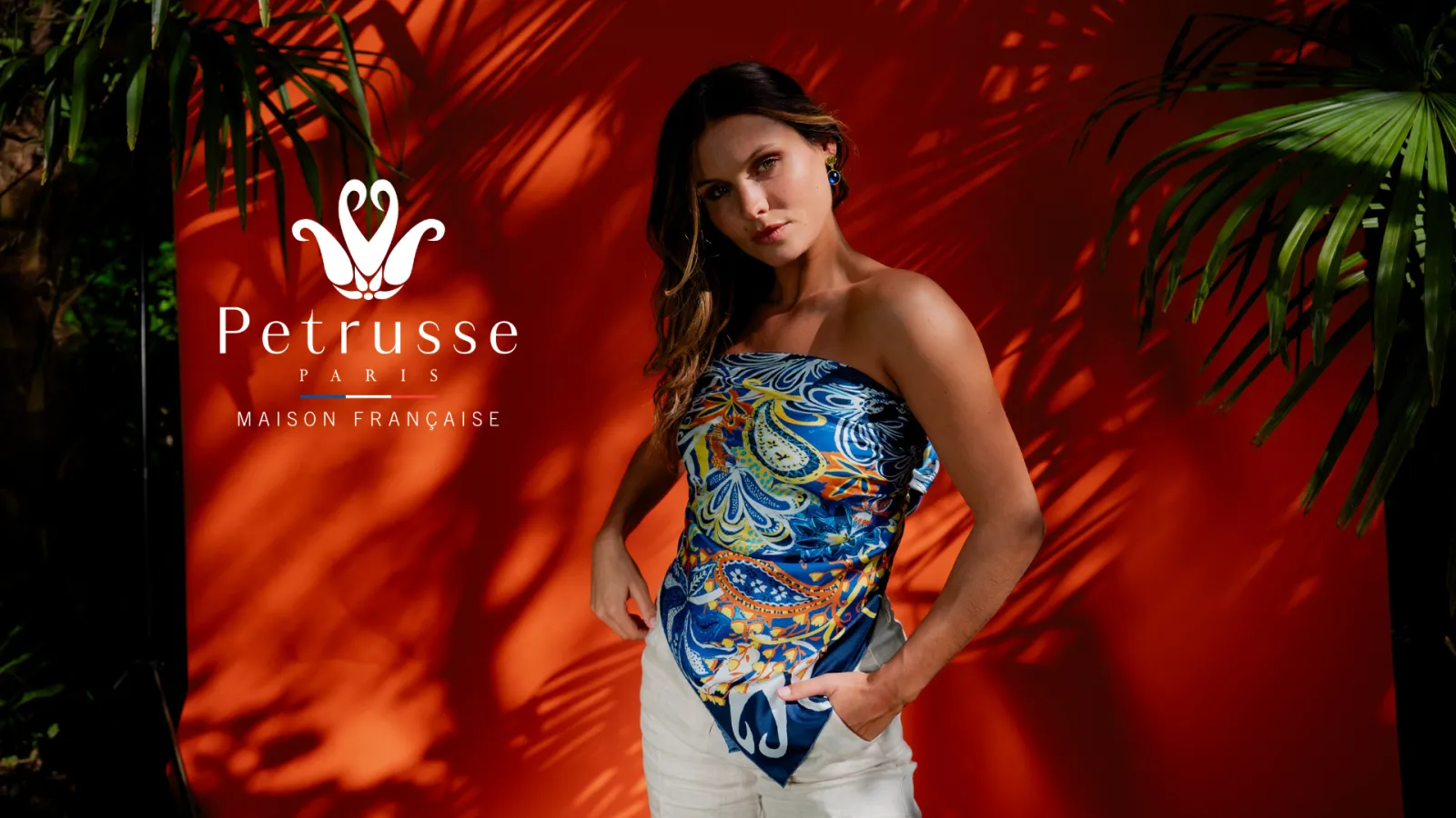 Woman wearing a blue strapless top with gold and white geometric patterns, posing against a red background with Petrusse Paris logo, French maison, with green plants in the background.