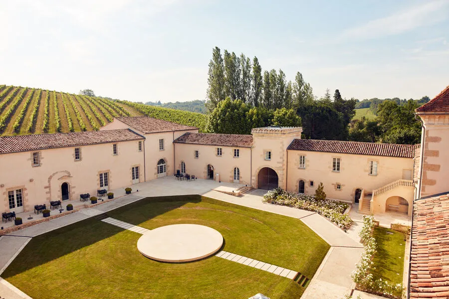 Aerial view of Château Toulouse-Lautrec near Bordeaux, featuring an interior courtyard with green lawn and white circular pool, surrounded by pink stone buildings with terracotta roofs, vineyards visible in the background.