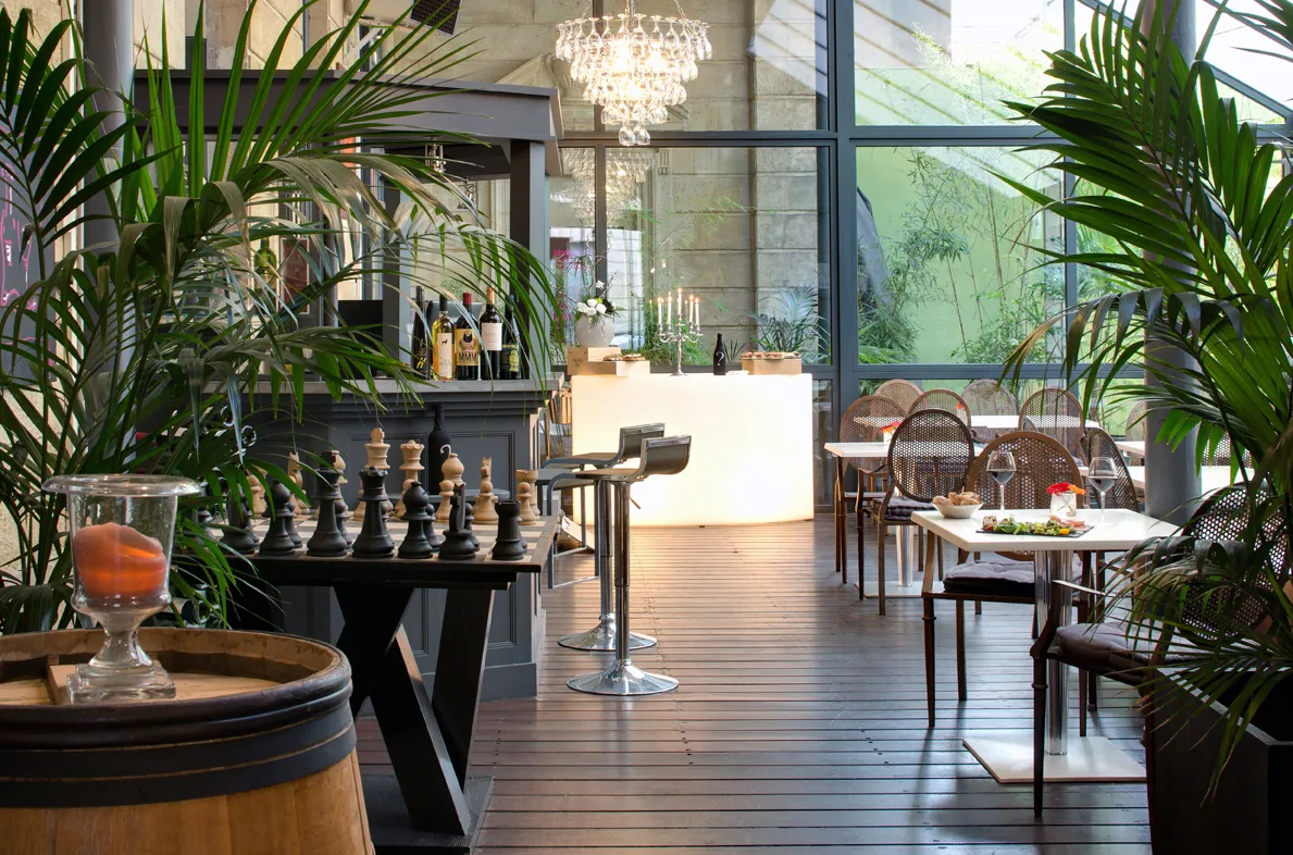Elegant interior of Bordeaux's Boutique Hotel Bar featuring a white bar counter, dining tables, woven rattan chairs, lush green plants, crystal chandelier and large windows overlooking a terrace and garden.