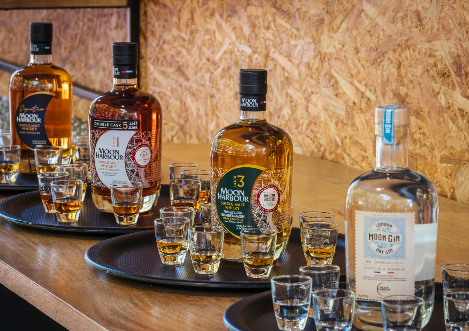 Moon Harbour spirits tasting in Bordeaux featuring multiple bottles of whisky and gin arranged on black trays with filled shot glasses against a cork wall backdrop.