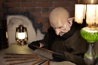 Bald fantastical character in black clothing examining a map with a magnifying glass in a vintage Bordeaux setting, surrounded by lanterns and mysterious objects.