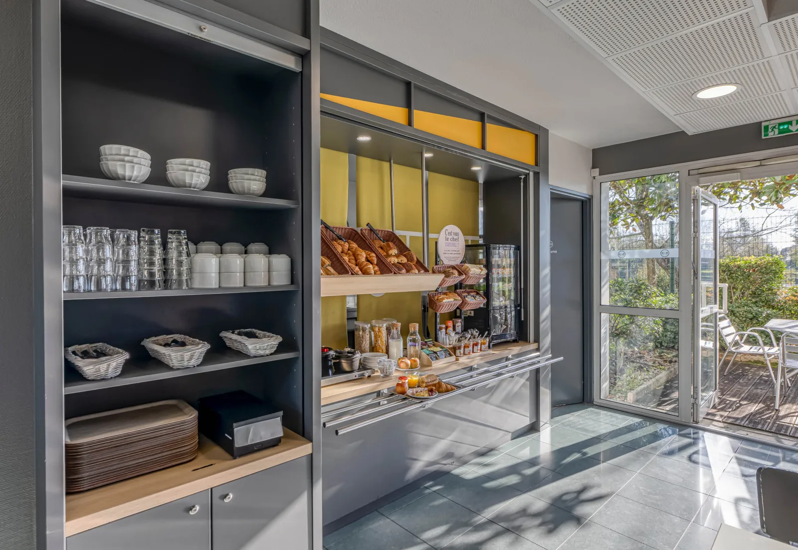 Breakfast area at B&B Hotel Bordeaux Bruges featuring storage shelves, glasses, bowls, baskets, display of pastries and breakfast items behind yellow glass window.