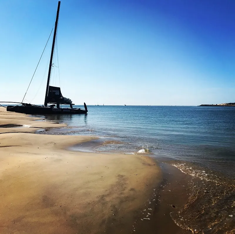 A black sailing yacht anchored near the shore on a sandy beach with turquoise sea and clear blue sky in the background, representing the Black Pearl of Arguin Sailing near Bordeaux.