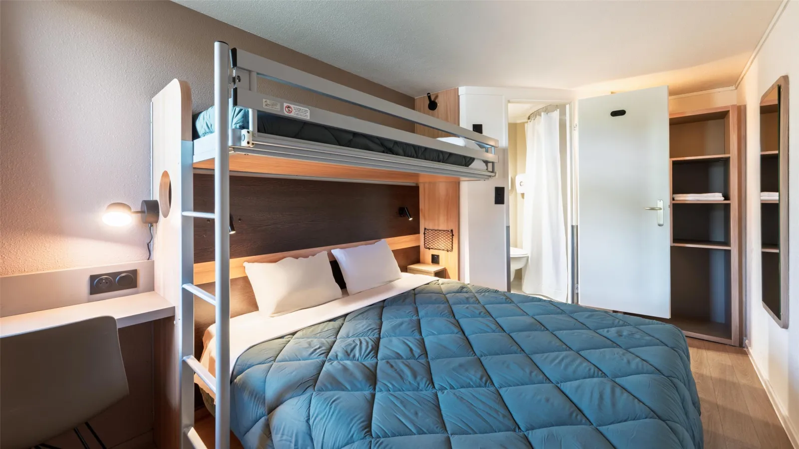 Modern hotel room featuring a metal and white bunk bed with blue quilt, white bedding, integrated desk with lamp, white wardrobe, visible bathroom, light wooden flooring and warm ambient lighting.