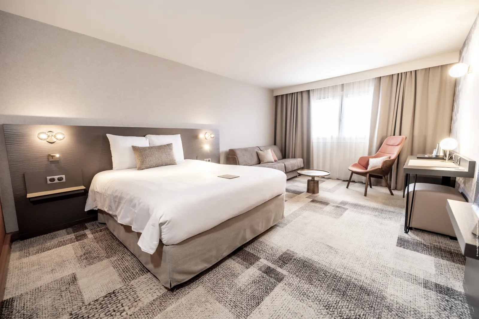 Modern hotel room at Mercure Bordeaux-Lac featuring a king-size bed, grey upholstered headboard with wall sconces, gray sofa, pink accent chair, large windows with beige curtains, contemporary furnishings, and neutral color scheme.