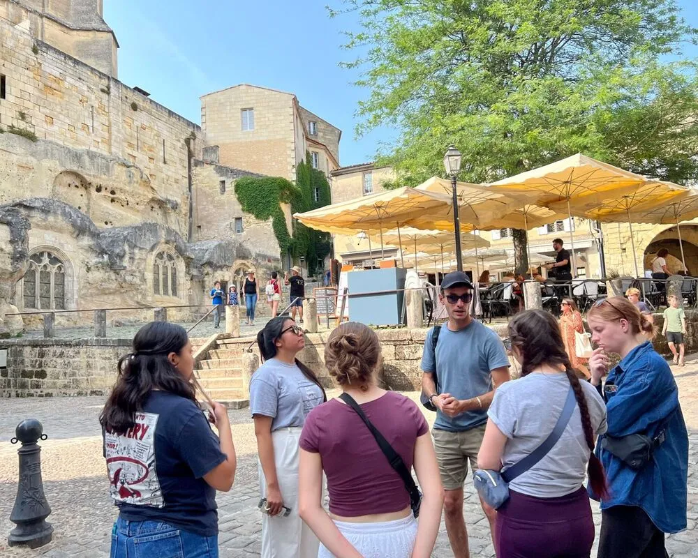 A group of tourists listening to a tour guide in front of a historic church in Bordeaux, sheltered under yellow umbrellas, with stone buildings and green trees in the background