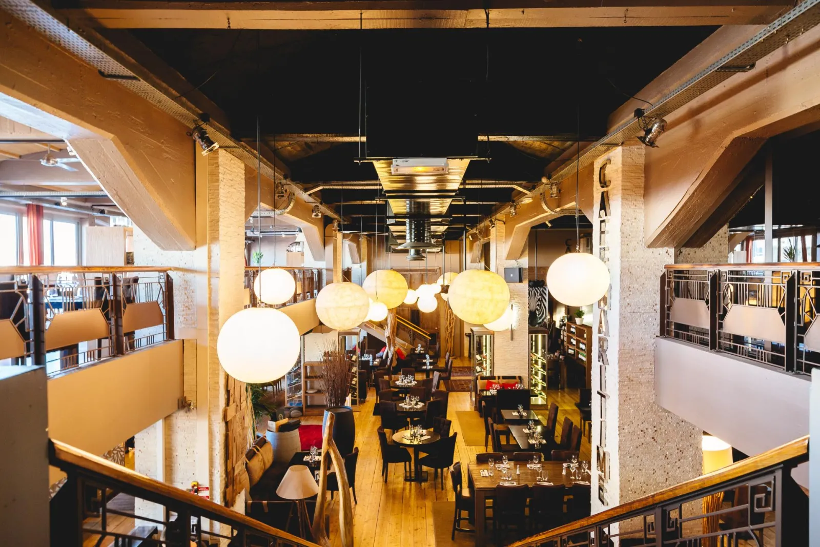 Interior view of Café Maritime in Bordeaux, featuring a modern restaurant with wooden beams, suspended spherical lights, black tables and wooden chairs, side galleries, and warm evening lighting.