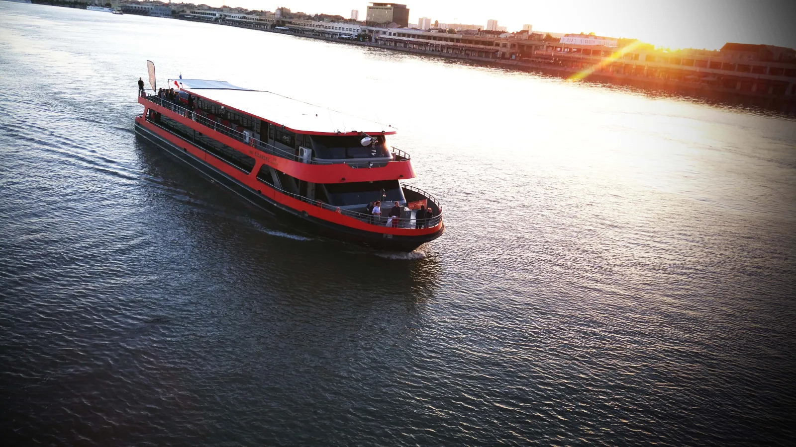 Red and black tourist boat sailing on the Garonne River in Bordeaux, with waterfront buildings and city skyline in the background at sunset
