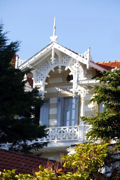 White Belle Époque villa with ornate wooden lace details, mansard roof and pointed turret, surrounded by greenery, in Arcachon's Ville d'Hiver
