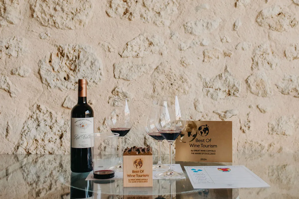 Red wine bottle from Château Rol Valentin with filled tasting glasses, Best Of Wine Tourism 2025 awards and presentation documents on white table against textured beige stone wall