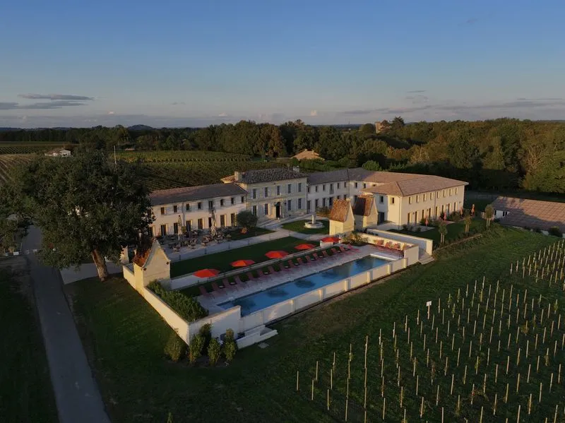 Aerial view of Château Fage, a luxury hotel-restaurant with ochre stone buildings, blue swimming pool, red loungers on terrace, surrounded by vineyards and forests in the Bordeaux region.