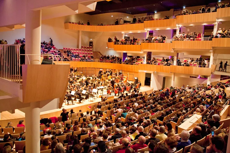 Interior view of the Bordeaux Auditorium featuring an orchestra performing on stage with musicians and instruments, surrounded by spectators seated in multi-level tiered seating. Modern architecture with light wood balconies and purple and orange ambient 