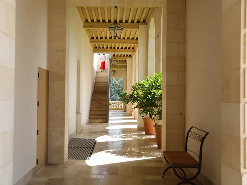 Covered gallery of Château Marquis d'Alesme Becker in Bordeaux, featuring white columns, wooden slatted ceiling, staircase in the background, and a potted plant.