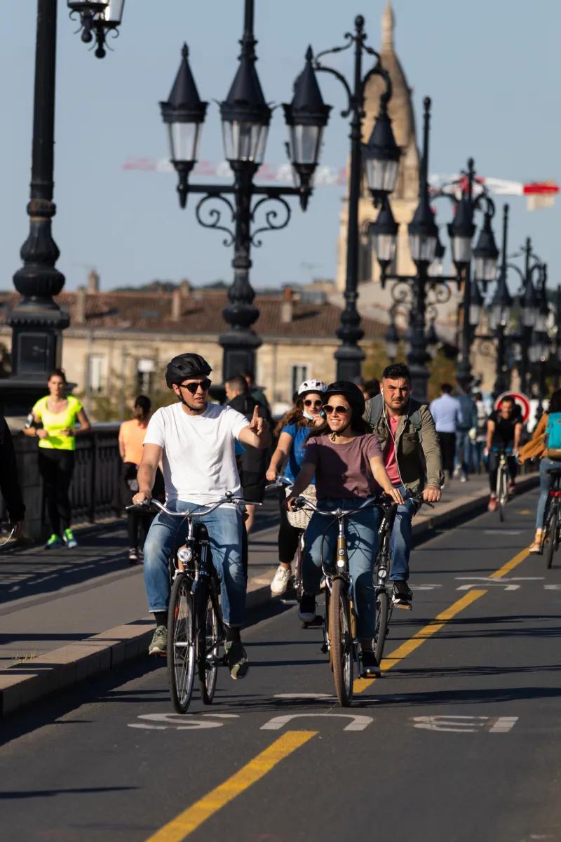 Cyclists riding bikes on the Stone Bridge in Bordeaux, with Saint-Michel Church in the background and distinctive black street lamps