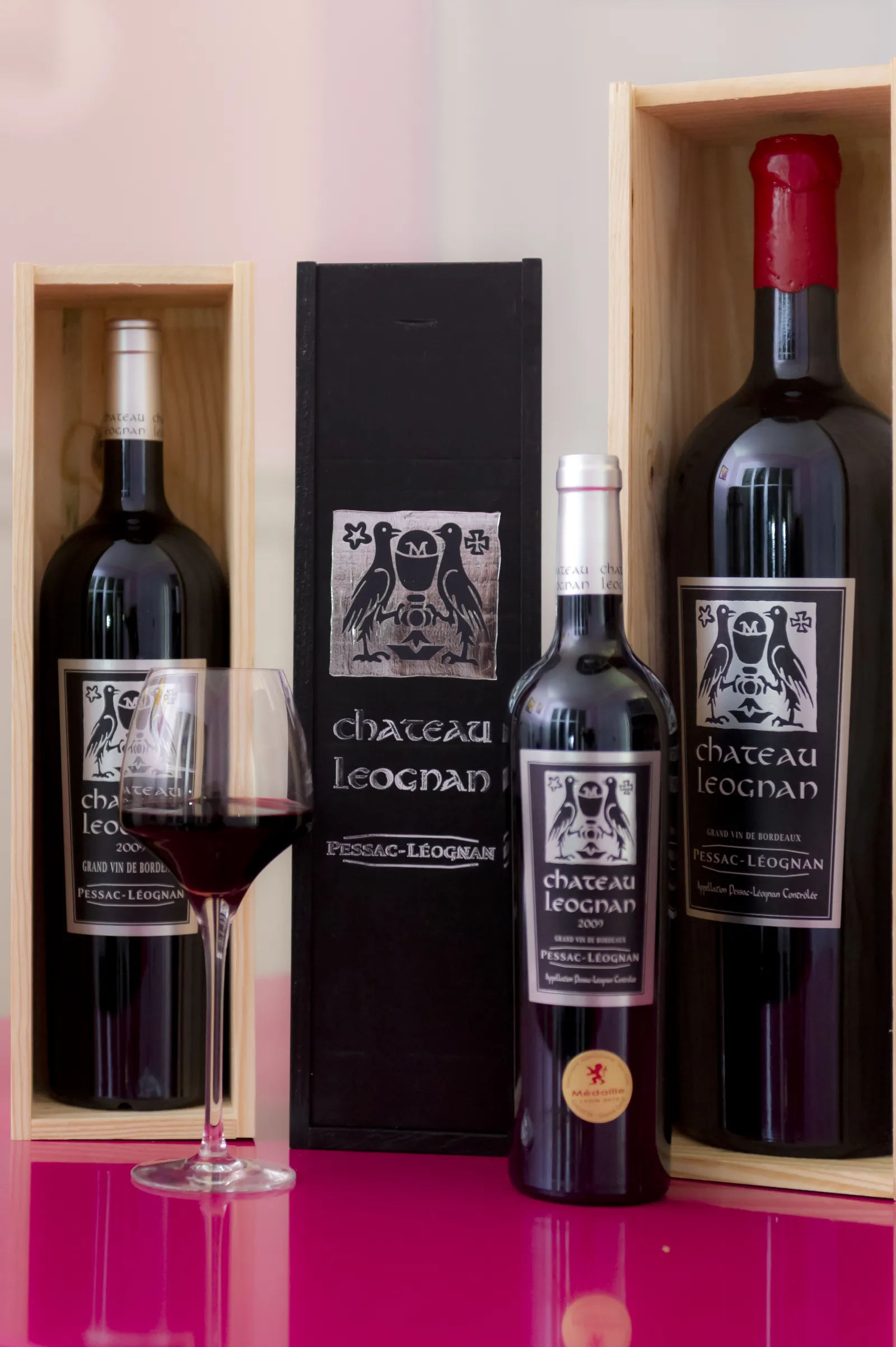 Elegant display of Château Léognan Bordeaux wines featuring bottles in wooden boxes, a wine glass, and black packaging on a pink background.