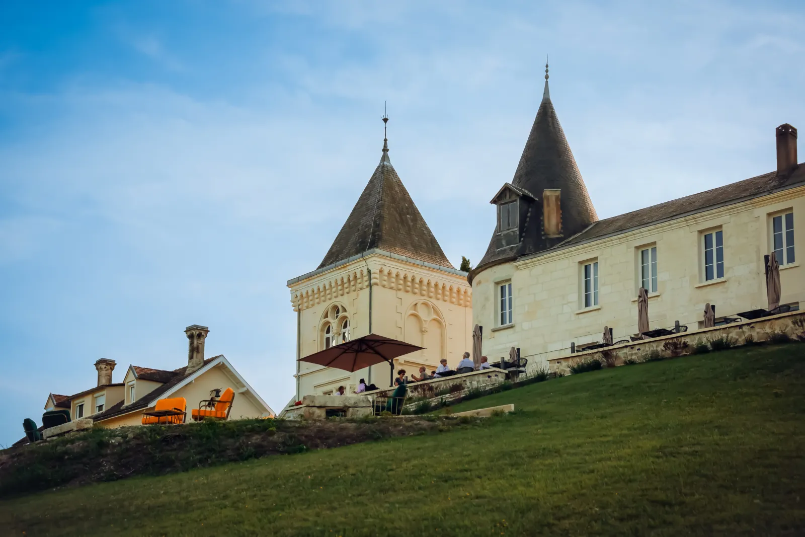 CarlMagnus Castle near Bordeaux, Roux Oulié house featuring distinctive conical towers, pale yellow facade, shaded terrace with orange chairs overlooking green grounds.