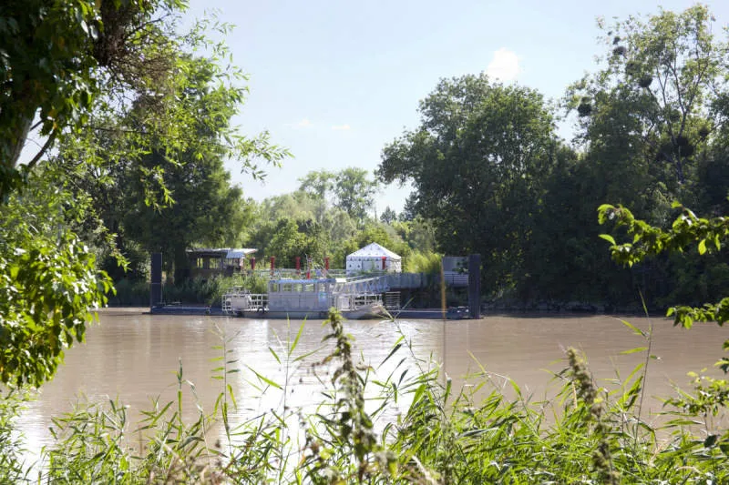 View of Arcins Island in Bordeaux with the Garonne river in the foreground, trees on both sides, and buildings visible on the island including a white tent.