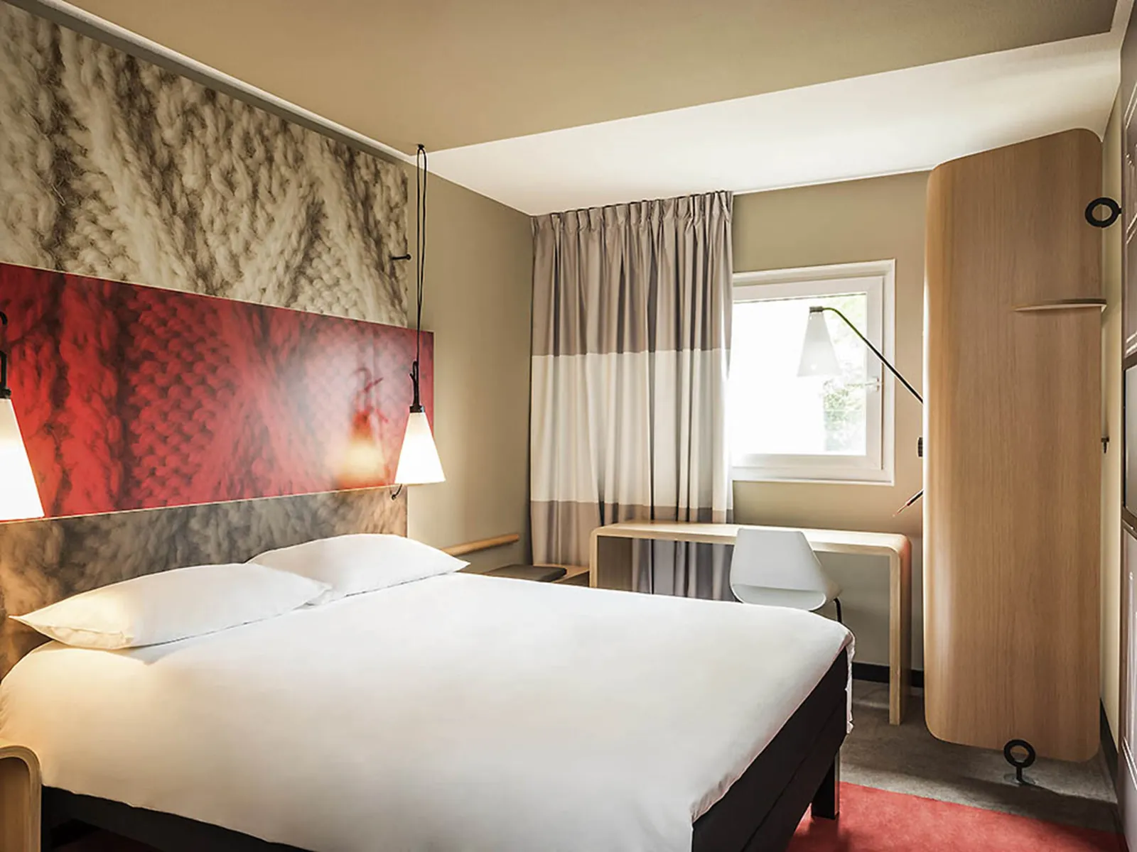Modern hotel room at Ibis Bordeaux Centre Bastide featuring a double bed with white linens, a colorful red and beige headboard, a window with gray and beige curtains, a light wood desk, and minimalist décor.