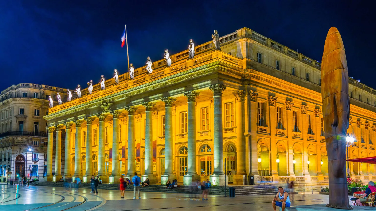 Bordeaux Grand Theatre lit up at night, yellow classical facade with columns, statues on the roof, French flag, paved square with visitors