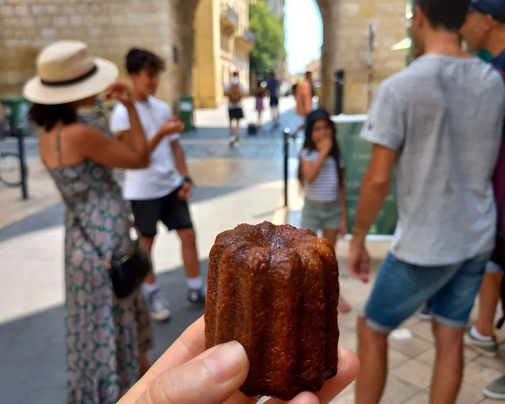 Hand holding a Bordeaux canelé, traditional pastry, in a cobblestone street of Bordeaux with tourists in the background