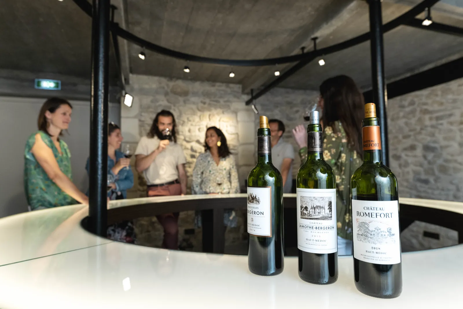 Three bottles of red wine from Château Romefort and Château Lamothe Bergeron displayed on a white counter, with visitors in the background at a Bordeaux wine tasting cellar.