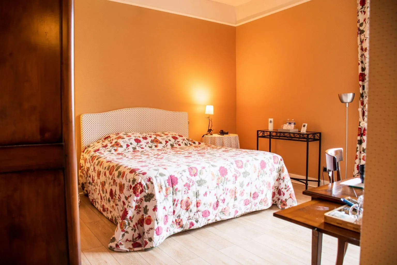 Hotel room with orange walls, queen-size bed with floral patterned bedspread, upholstered headboard, nightstand with lamp, mirror, desk and floral curtains at Les Chambres d'Arche in Bordeaux