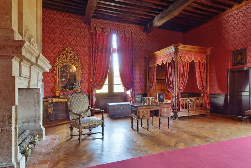 Royal bedroom at Château de Cazeneuve featuring damask red walls, period furniture, ornate fireplace, gilded canopy bed, and burgundy draped windows.