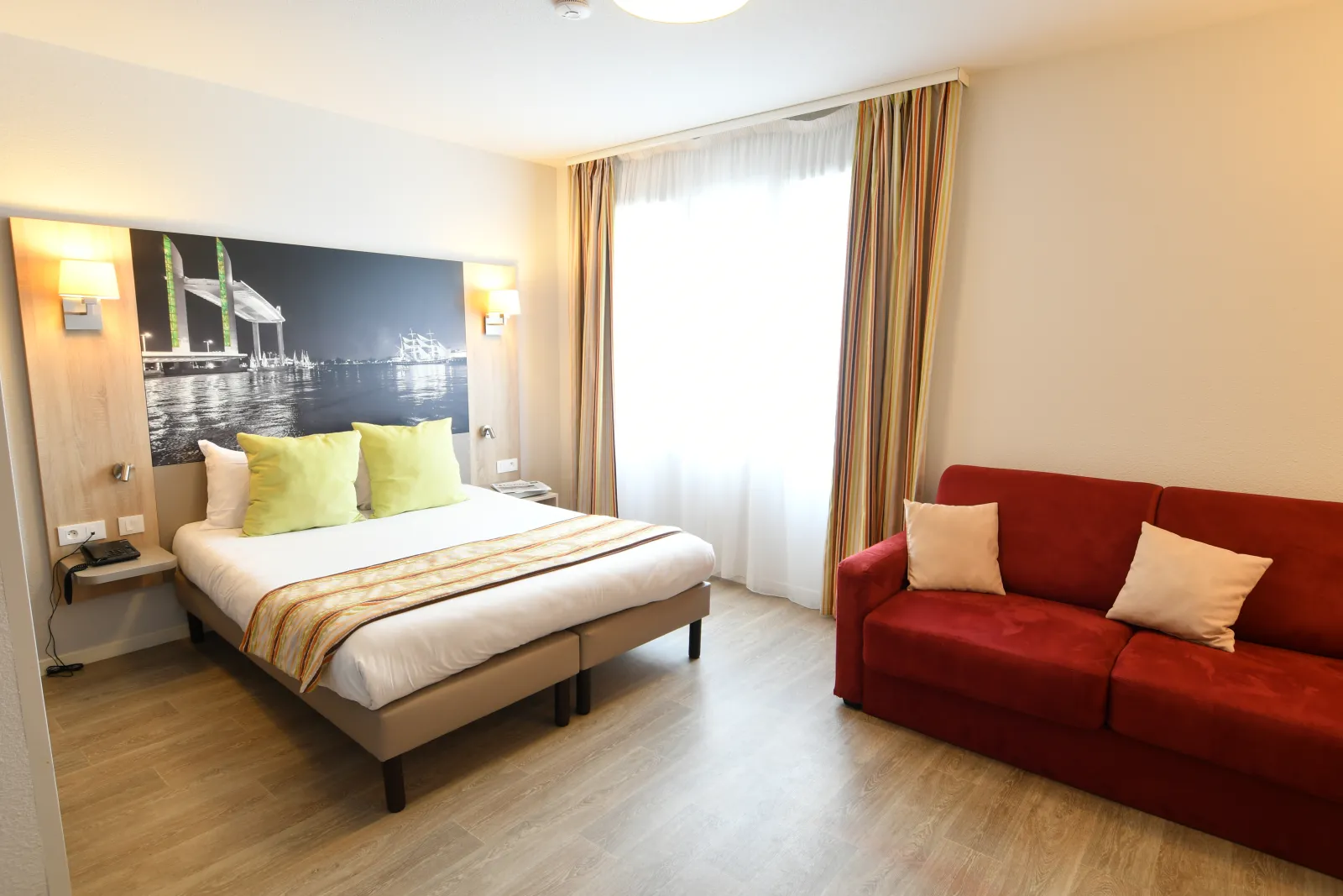 Spacious hotel room in Bordeaux featuring a double bed with yellow cushions, headboard with illuminated bridge photograph, red sofa with beige pillows, large windows with gold curtains, light wood flooring, and wall sconces.