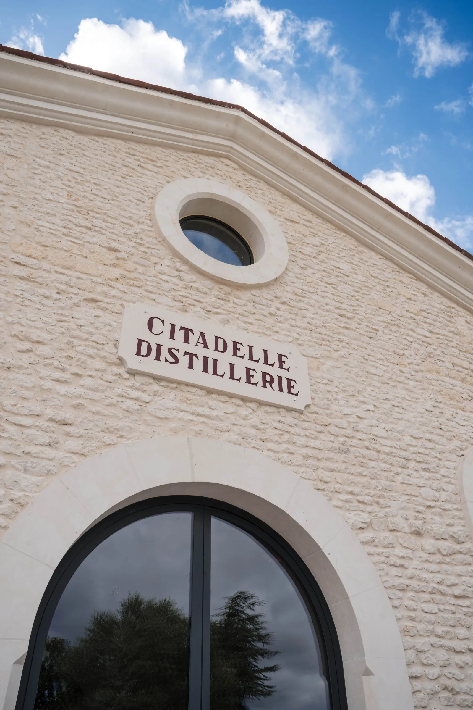 White stone facade of Citadelle Distillery in Bordeaux featuring an engraved sign, circular window, and large glass window