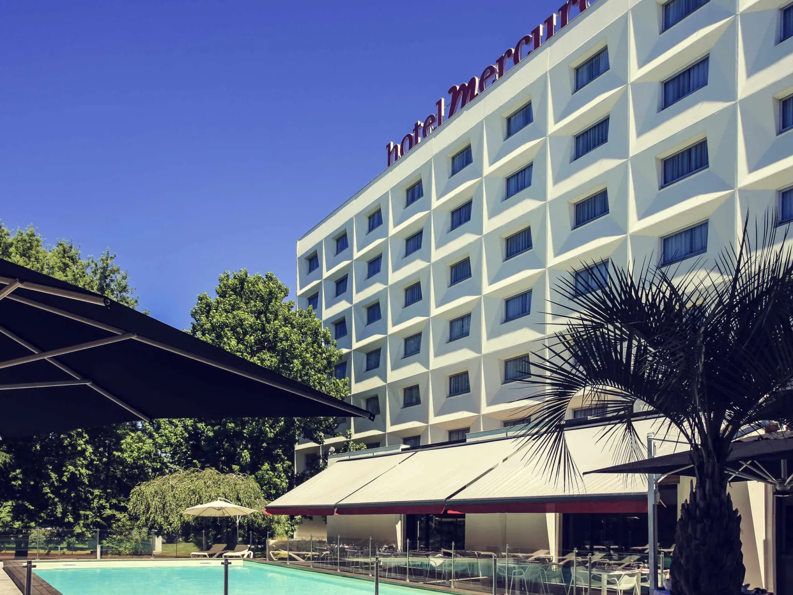 Modern white hotel building with swimming pool and shaded terrace overlooking Bordeaux lake, surrounded by palm trees and green spaces
